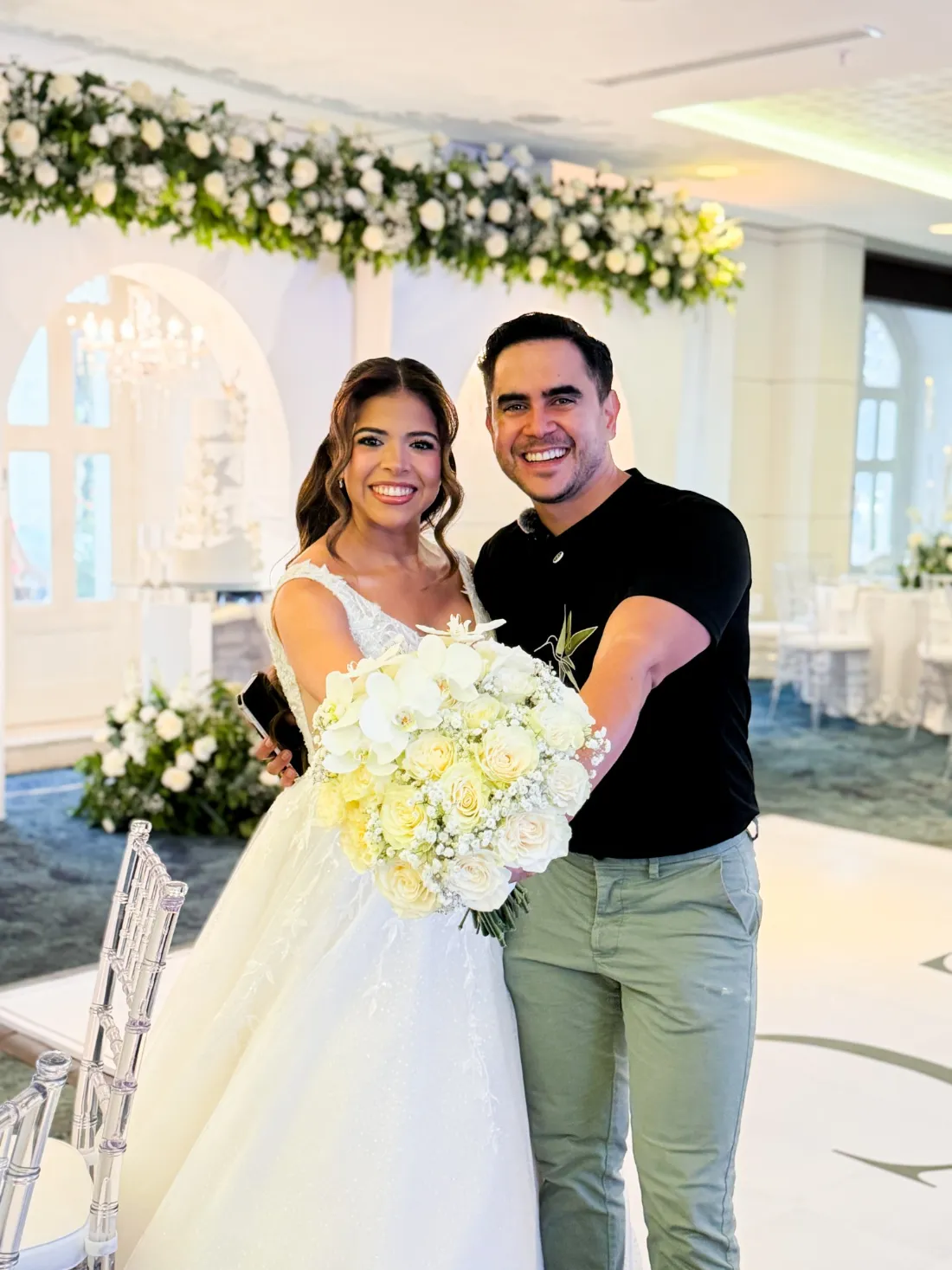 Andy Colmenares y una novia con ramo de rosas blancas y orquídeas bajo una guirnalda floral