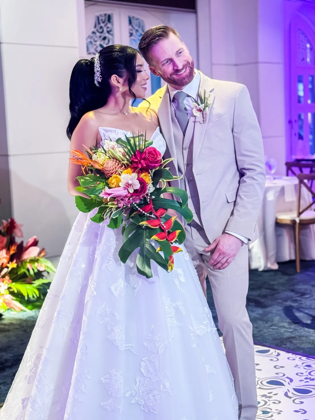 Novios con ramo en cascada de heliconias, orquídeas y rosas sobre hojas de monstera