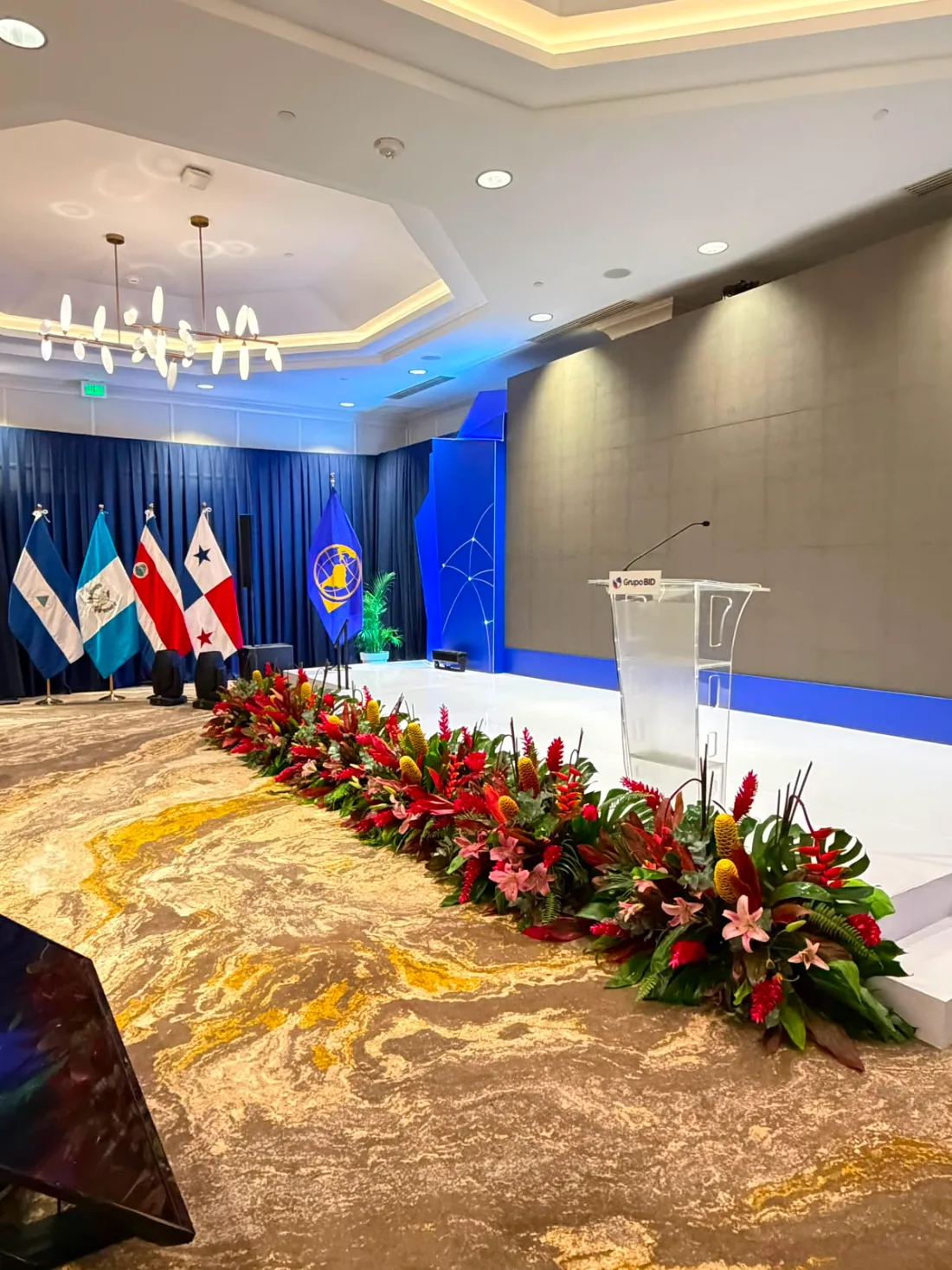 Escenario de evento corporativo con atril, banderas y arreglos florales tropicales