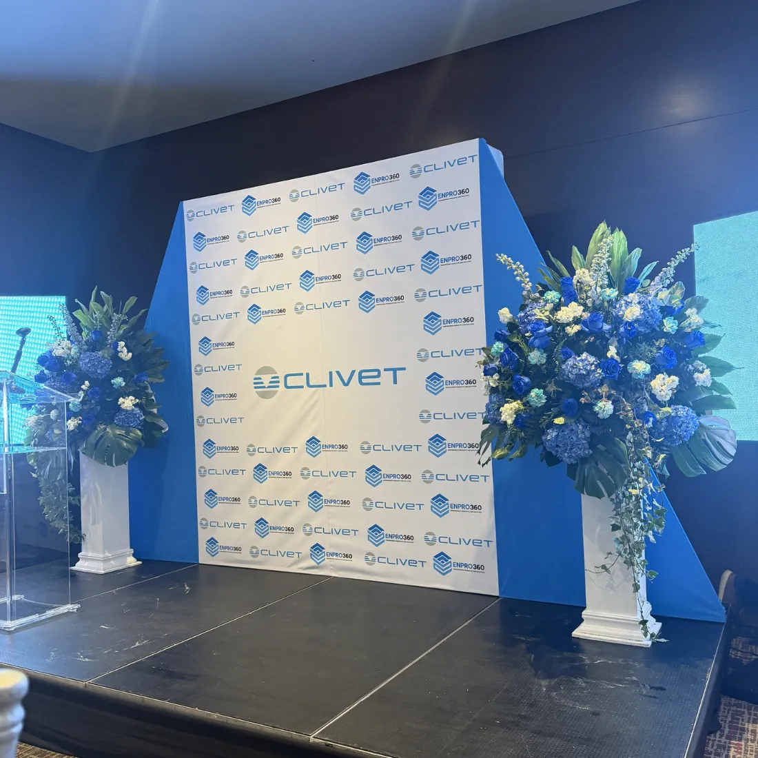 Escenario corporativo con backing de marca y arreglos florales azules